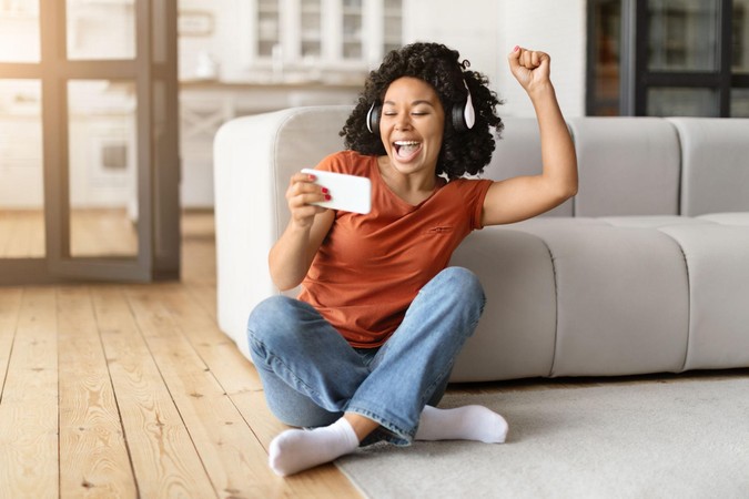 Mwanamke mwenye furaha amekaa sakafuni akitumia simu na kusherehekea ushindi.