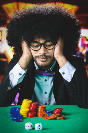 Mwanaume mwenye nywele nyingi na miwani amekaa mbele ya chips za kasino akiwa na wasiwasi.