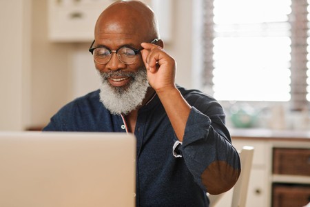 Mwanaume mwenye ndevu nyeupe na miwani anatabasamu huku akitazama laptop mezani nyumbani.