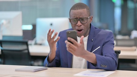 Mwanaume ofisini anaangalia simu kwa mshangao akiwa ameinua mkono mmoja.