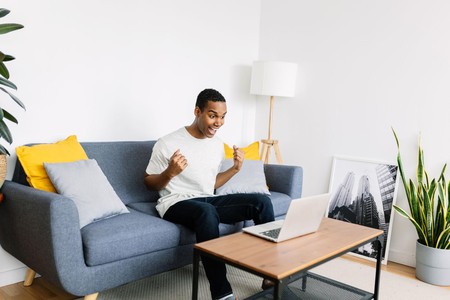 Kijana amekaa kwenye sofa mbele ya laptop akishangilia kwa furaha nyumbani.