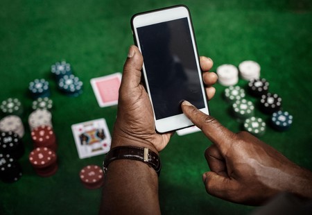 Mkono wa mtu ukitumia simu juu ya meza ya mchezo yenye chipsi za poker na kadi.