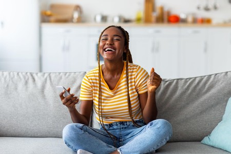 Msichana mwenye furaha ameketi kwenye sofa akiwa ameshika simu nyumbani.