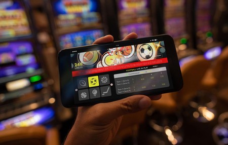 Simu inaonyesha programu ya kubashiri michezo huku mashine za slot zikionekana nyuma.