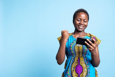 Mwanamke mwenye tabasamu anaangalia simu na kushangilia kwa mkono mmoja juu ya mandharinyuma ya bluu.
