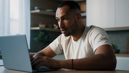 Mwanaume anatazama laptop kwa umakini akiwa nyumbani kana kwamba anafuatilia matokeo mtandaoni.