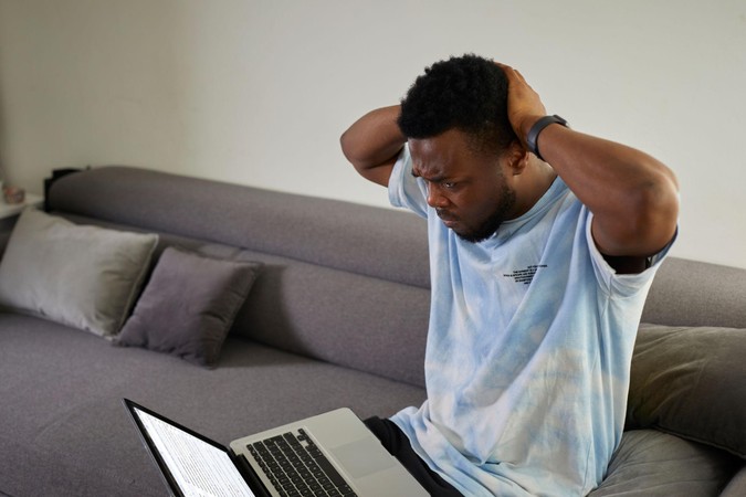 Mwanaume amekaa kwenye sofa akiwa ameshika kichwa huku akitazama laptop, akionyesha msongo au kukatishwa tamaa baada ya kucheza mtandaoni.