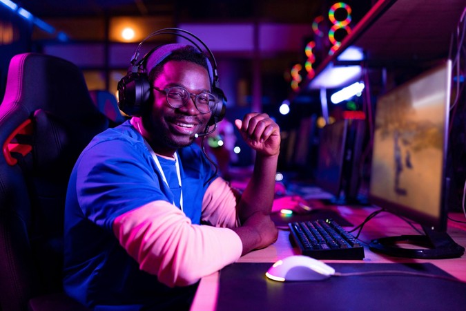 Mchezaji mwenye headset anatabasamu akiwa mbele ya kompyuta kwenye chumba cha michezo chenye taa za rangi, akionyesha burudani ya gaming.