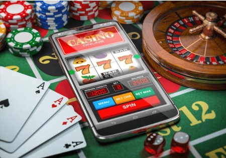 Simu yenye mchezo wa casino mtandaoni imewekwa juu ya meza ya kamari karibu na kadi, kete, chips na gurudumu la roulette.
