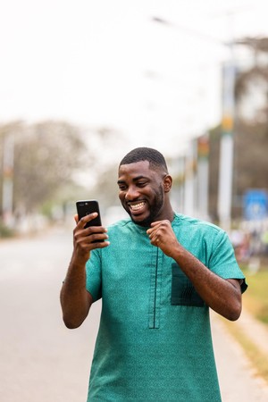 Mwanaume anatabasamu akiwa nje barabarani huku akiangalia simu yake na kushangilia kana kwamba amepokea matokeo mazuri.