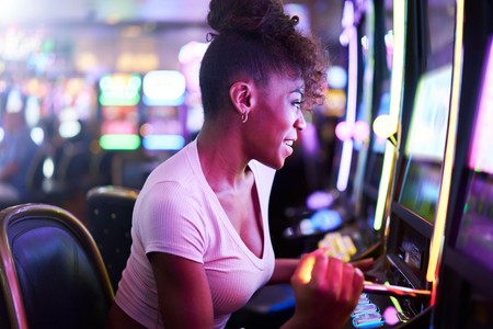 Mwanamke ameketi mbele ya mashine ya slot kwenye ukumbi wa casino wenye taa za rangi na anaonekana kufurahia mchezo.