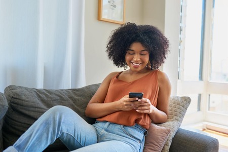 Mwanamke ameketi kwenye sofa akiwa ametulia na kutabasamu huku akitumia simu yake.