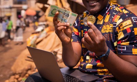 Mwanaume ameketi nje na laptop akionyesha noti ya fedha na sarafu ya Bitcoin mkononi.